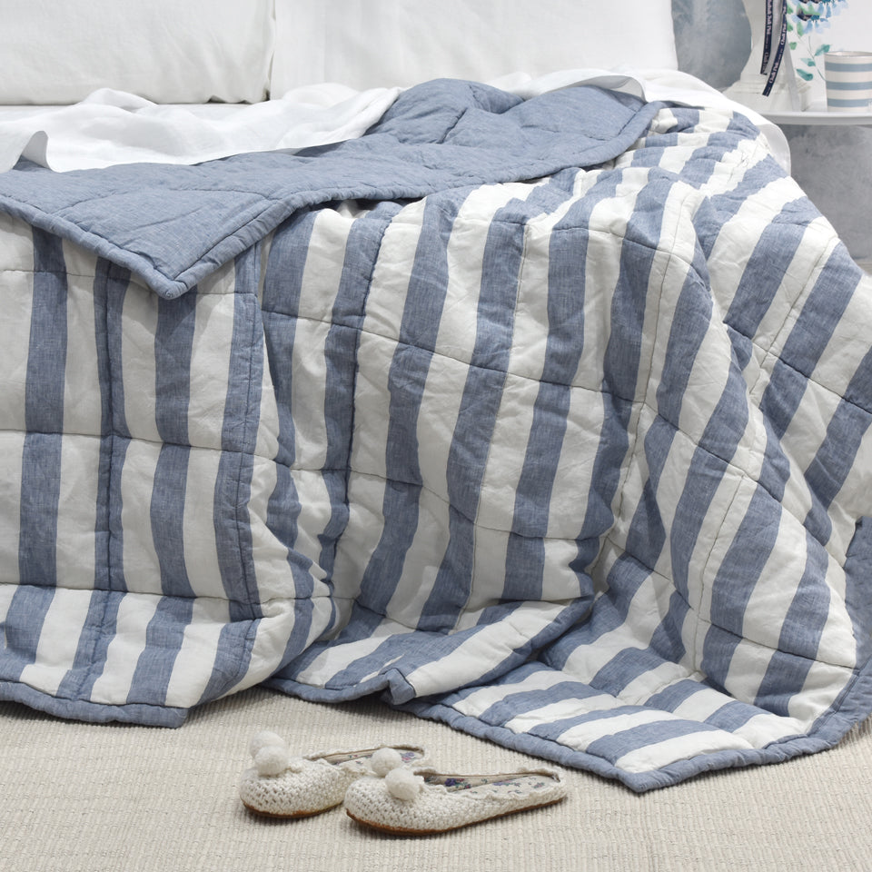 A Blue Chambray Stripe Linen Quilted Bedspread on a Bed with White Linen Sheets and Cream Slippers at the side of the bed