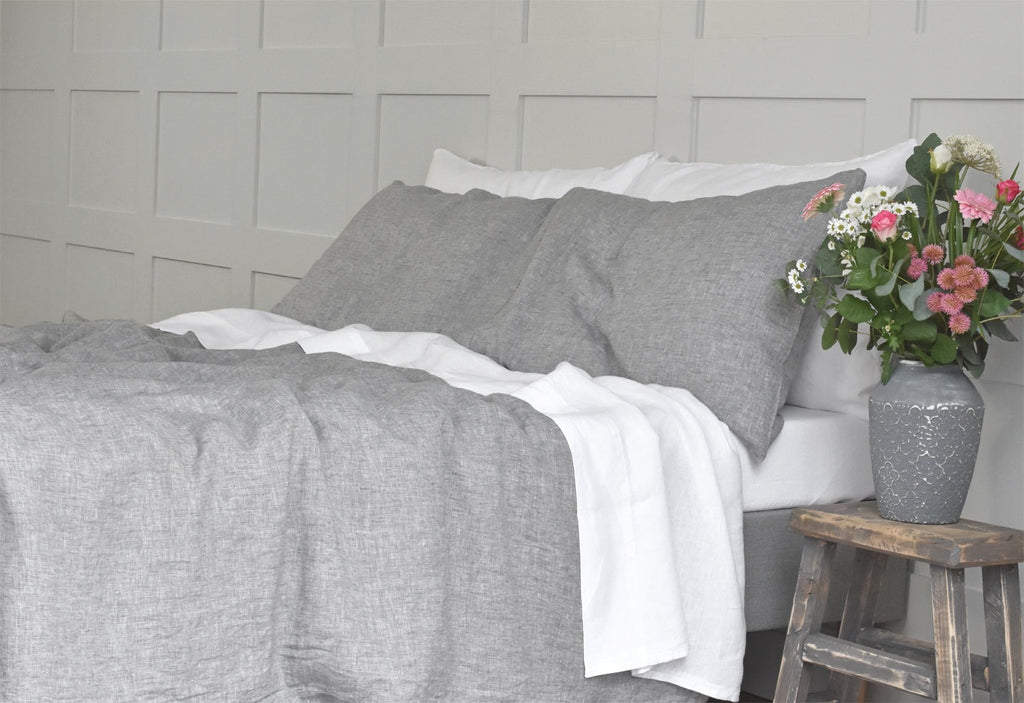 A Bed with Grey Linen Bedding and a White Linen Sheet with a side table and Pink Flowers