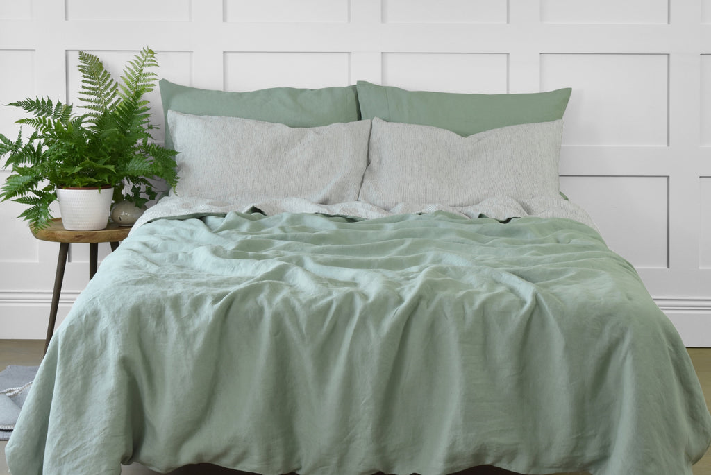 A Sage Green Linen Duvet Cover with Pinstripe Linen Backing, on a Bed with Stripe Linen Pillowcases and a side table with a big green fern plant