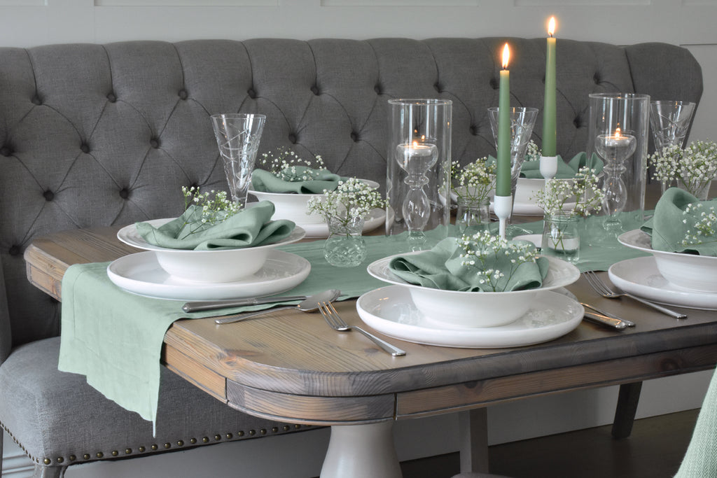 A Sage Green Linen Table Runner on a Table with White Flowers, and White Crockery with Green Linen Napkins in Bowls