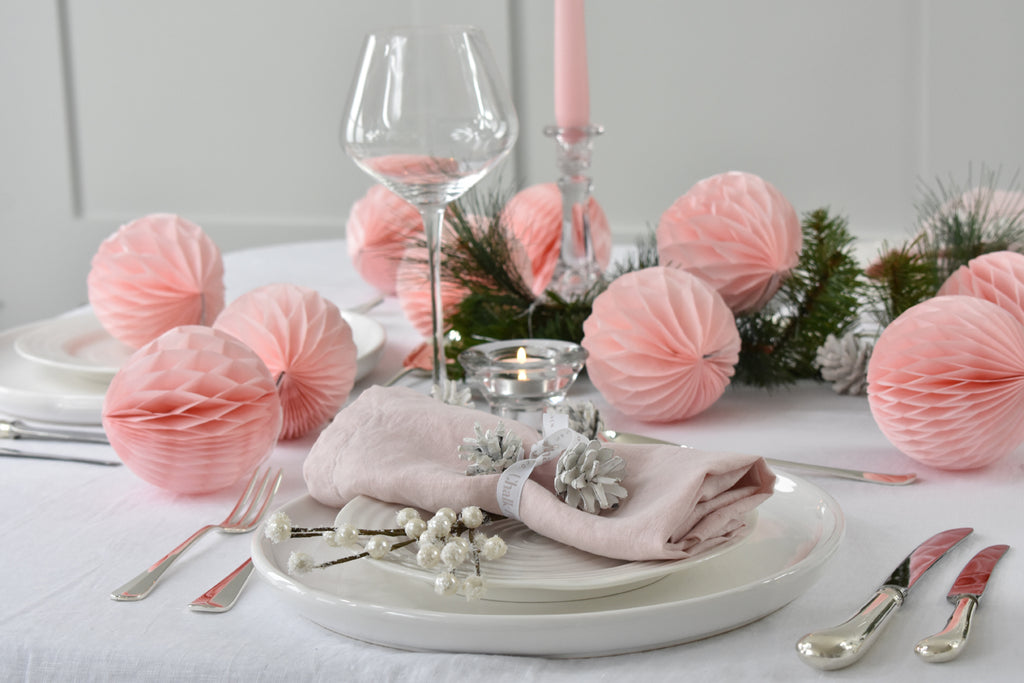 Decorative table setting with pink paper balls, candles, and greenery on a white tablecloth.