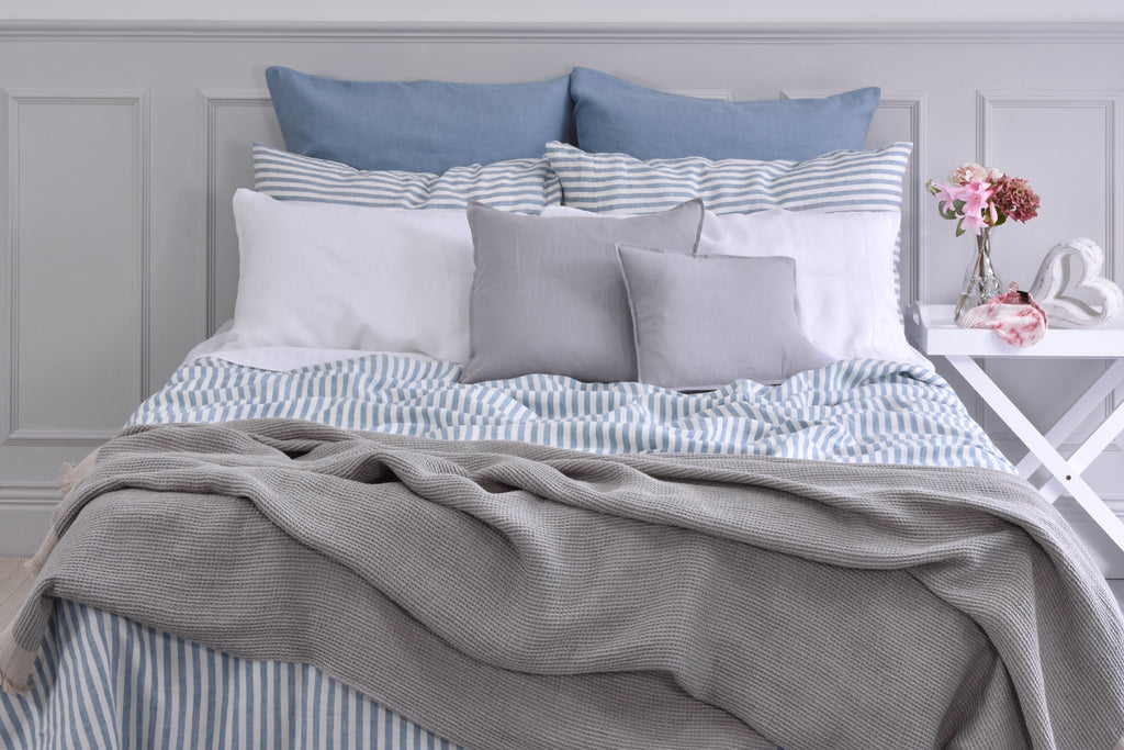 A Bed with Blue Stripe Linen Bedding and Grey Linen Cushions with a Grey Linen Blanket and Pink Flowers