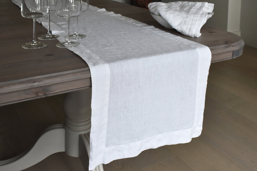A White Linen Table Runner on a Brown Oak Table with White Linen Napkins and Wine Glasses